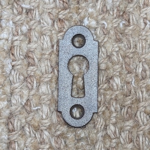 May include: A silver keyhole cover with a rounded top and bottom. The cover has a keyhole shape cut out in the centre.