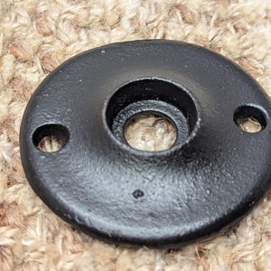 May include: Black metal pipe flange with two holes and a large center hole. The flange is smooth and has a matte finish.