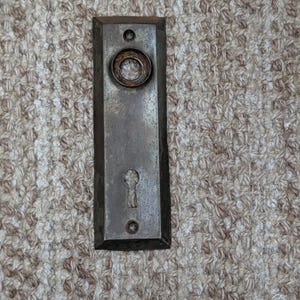 Pode incluir: Uma placa de porta retangular de metal desgastado com um acabamento escuro e manchado. A placa apresenta uma abertura circular no topo para o puxador e uma fechadura abaixo, com pequenos orifícios para parafusos nas extremidades.
