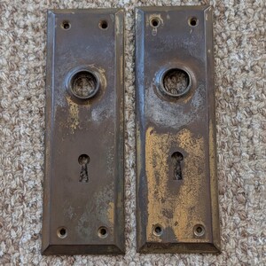 May include: Two rectangular brass door plates with a weathered, tarnished finish. Each plate features a central circular opening for a door knob, a lower keyhole opening, and four small screw holes located at the top and bottom corners.