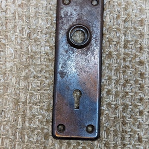 Peut inclure: Une plaque de porte rectangulaire en métal vintage avec un trou de serrure et un trou de bouton rond. La plaque est brun foncé avec une finition usée et vieillie.