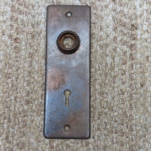 Peut inclure: Une plaque de porte rectangulaire en métal vintage avec un trou de serrure et un bouton rond en métal. La plaque est rouillée et a un aspect usé et vieilli.