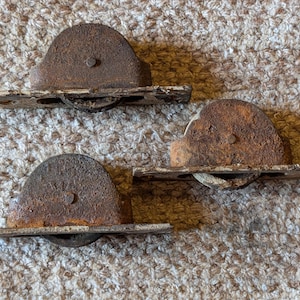 Könnte beinhalten: Drei antike, rostige Metall-Türrollen. Jede Rolle hat eine gebogene, halbkreisförmige Oberseite mit einem kleinen Metallstift und eine rechteckige Basis mit einem Rad. Die Rollen sind stark verwittert mit einem braunen und weißen Farbschema.