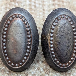 May include: Two oval-shaped, black metal drawer pulls with a beaded border and a slightly raised center. The pulls have a distressed, aged finish.