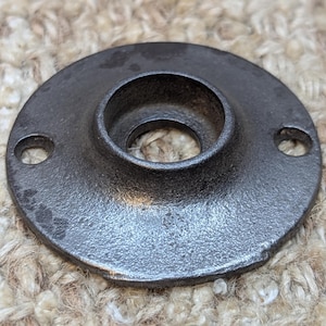 May include: A black metal pipe flange with a round opening and two screw holes. The flange has a rough, textured surface.
