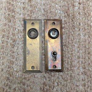 Puede incluir: Dos placas de puerta rectangulares de latón con bordes biselados. La placa izquierda presenta una abertura circular central y dos orificios para tornillos. La placa derecha incluye una abertura circular, un pequeño mecanismo de botón vertical.