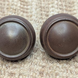 May include: Two round, dark brown cabinet knobs with a raised, circular center. The knobs have a slightly textured surface.