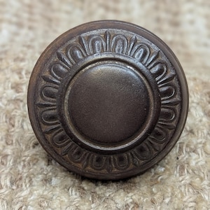 May include: A dark brown round door knob with a decorative, raised, circular design around the edge.