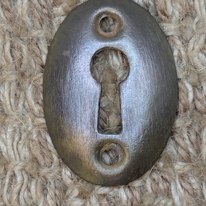 May include: A vintage oval-shaped keyhole cover made of metal with a brushed finish. The cover is a dark grey colour with a slightly greenish hue.