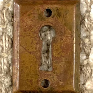 Könnte beinhalten: Ein rechteckiger Schlüsselschlitzabdeckung aus Messing mit einem Schlüsselschlitz in der Mitte und zwei kleinen Löchern an beiden Seiten. Die Abdeckung hat ein rustikales, gealtertes Aussehen.