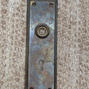 Pode incluir: Uma placa de porta retangular de metal com acabamento desgastado e manchado, apresentando uma abertura circular central para maçaneta e uma fechadura inferior. A placa possui quatro pequenos orifícios para parafusos e oxidação.
