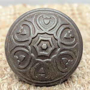 May include: A round, dark gray metal door knob with an intricate, embossed design. The design features a central starburst surrounded by six stylized floral motifs.