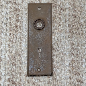 Pode incluir: Uma placa de porta retangular de metal castanho desgastado, com uma abertura circular para puxador no topo e uma fechadura vertical na parte inferior. A placa inclui dois pequenos orifícios circulares para parafusos, um no topo e outro na base.