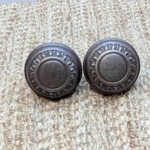 May include: Two round, dark gray metal cabinet knobs with a decorative, raised, circular design around the edge.