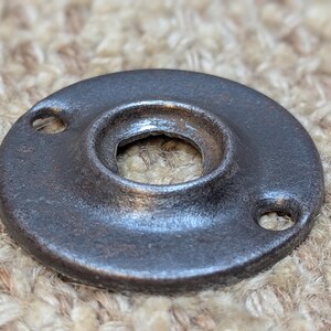 May include: A round, dark gray metal plate with a hole in the center and two smaller holes on the sides. The plate has a slightly textured surface.