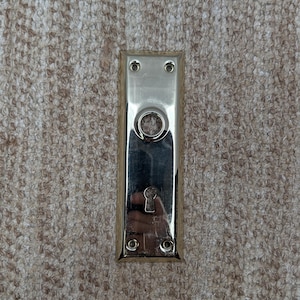 Puede incluir: Una placa de puerta rectangular de latón pulido que presenta una abertura circular central para un pomo y una abertura inferior para una cerradura. La placa incluye cuatro pequeños orificios para tornillos para su montaje en una puerta.