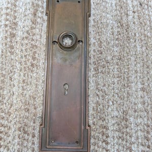 Op de afbeelding: Een rechthoekige bronzen deurplaat met een verweerde patina, voorzien van een centrale ronde opening voor een deurknop en een sleutelgat eronder. De plaat heeft een getrapte rand en vier kleine schroefgaten in de hoeken.