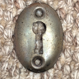 Op de afbeelding: Ovale, antieke metalen sleutelplaat met een sleutelgat. De plaat heeft een aangetaste, verouderde uitstraling met twee kleine klinknagels boven en onder het sleutelgat. De achtergrond is een textiel met neutrale tinten.