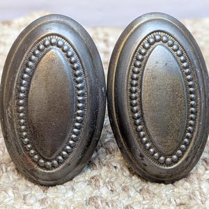 May include: Two antique oval cabinet knobs with a dark bronze finish. Each knob features a central oval design surrounded by a beaded border. The knobs are likely made of metal and show signs of age.