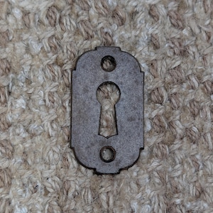 May include: A rectangular metal keyhole escutcheon plate with rounded edges and a central keyhole cutout. The plate features two circular screw holes, one positioned above and one below the central keyhole, and has a dark, weathered finish.