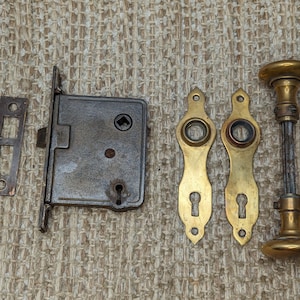 May include: Antique brass door hardware set with a door knob, two escutcheons, and a strike plate. The hardware is in good condition with some signs of wear.