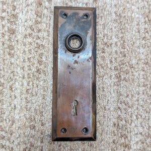 May include: A rectangular brass door plate with a weathered, tarnished finish. The plate features a central circular opening for a door knob and a lower keyhole opening. Four small screw holes are positioned at the top and bottom corners.
