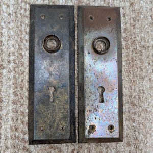 Pode incluir: Duas placas de porta retangulares de metal com um acabamento escuro e desgastado. Cada placa apresenta uma abertura circular central para uma maçaneta, uma fechadura abaixo e quatro pequenos orifícios para parafusos nos cantos.