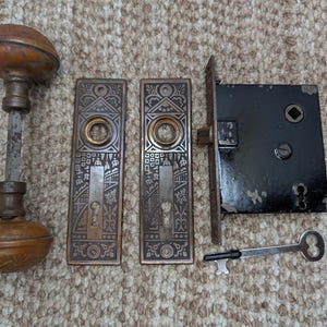 May include: Antique door hardware set including a bronze doorknob, two ornate bronze door plates, a black metal lock, and a silver key. The door plates feature intricate patterns and keyhole details. The set is likely from the early 20th century.