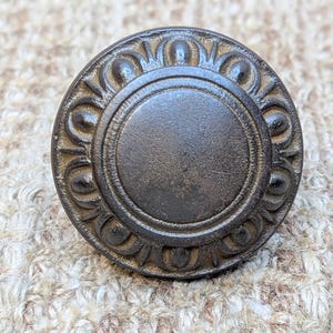 May include: A round, dark brown metal knob with a decorative, raised design around the edge. The knob has a smooth, slightly textured surface.