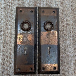 Könnte beinhalten: Zwei rechteckige Türschilder aus Metall mit verwitterter Bronzeoberfläche. Jedes Schild verfügt über eine kreisförmige Öffnung für den Türknauf oben und ein Schlüsselloch unten sowie vier kleine Schraublöcher an den Ecken.