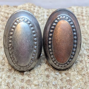 May include: Two oval-shaped metal drawer pulls with a decorative beaded border. One pull is silver and the other is copper.