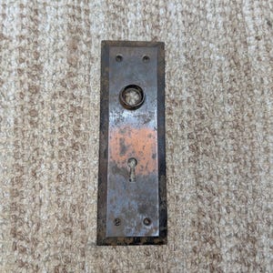 Pode incluir: Uma placa de porta retangular de metal com uma pátina escura e desgastada e uma seção central em tons de cobre. A placa apresenta uma abertura circular no topo para o puxador e um orifício clássico para a chave na parte inferior.