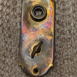 May include: An antique brass door plate with a tarnished, iridescent finish. The plate has a keyhole and a small, decorative handle. Two screw holes are visible at the top and bottom. The overall shape is rectangular with rounded edges.