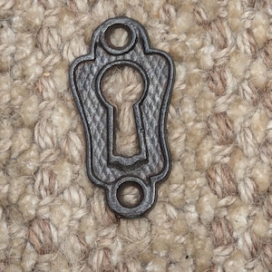 May include: A black metal keyhole cover with a decorative design. The cover has a round hole at the top and bottom.