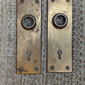 May include: Two antique brass door plates with keyholes and round decorative holes. The plates have a worn, aged finish.