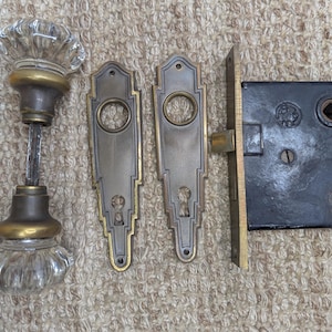 May include: Antique door hardware set featuring a clear glass doorknob with brass accents, two decorative backplates, and a black metal mortise lock. The hardware has an aged appearance, suggesting vintage or retro styling.