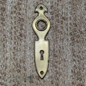 Puede incluir: Una placa de escudo para puerta vertical de latón con extremos puntiagudos, una abertura circular para el eje de la manilla y una cerradura inferior. La placa de metal incluye dos pequeños orificios circulares para tornillos de montaje.