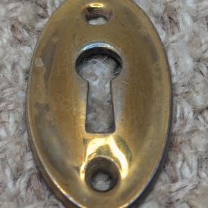 Op de afbeelding: Een messing sleutelgat afdekking in een ovale vorm met twee kleine gaten aan de onderkant. Het sleutelgat bevindt zich in het midden van de afdekking.