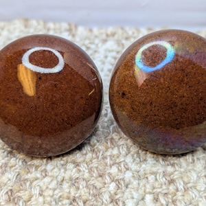 Pode incluir: Dois botões de cerâmica marrom com acabamento brilhante. Um botão tem um design de círculo branco, enquanto o outro tem um design de círculo azul e iridescente.