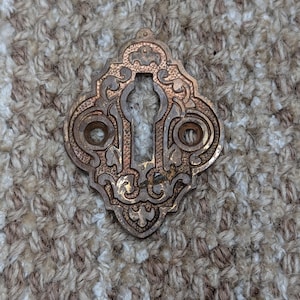 Könnte beinhalten: Verzierter Schlüsselloch-Abdeckung im antiken Stil mit Bronze-Finish. Die Abdeckung hat ein dekoratives, symmetrisches Design mit einem Schlüsselloch und zwei kreisförmigen Löchern. Das Metall hat ein verwittertes Aussehen, das auf Alter und Gebrauch hindeutet. Dekorationselement.