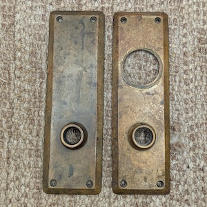 May include: Two antique brass door plates with a rectangular shape and rounded edges. The plates have a keyhole and a round knob hole.