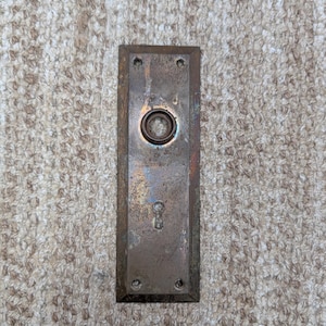 Op de afbeelding: Een rechthoekige, verweerde koperen deurplaat met een centrale ronde opening voor een deurknop en een sleutelgat eronder. Het metalen oppervlak vertoont een verouderde patina met verkleuringen en vier kleine schroefgaten op de hoeken.