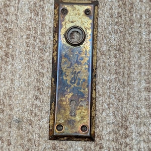 May include: A rectangular metal door plate featuring a weathered gold and dark patina finish. The plate includes a circular opening for a door knob at the top and a keyhole opening at the bottom, with four small screw holes at the corners.