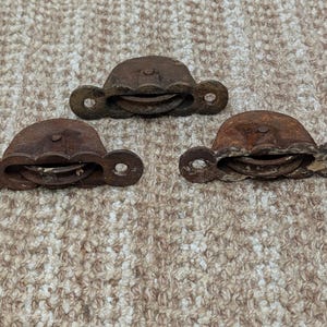 Könnte beinhalten: Drei antike, rostige Metallrollen. Jede Rolle hat eine halbkreisförmige Oberseite mit einem kleinen Rad in der Mitte und zwei kreisförmigen Löchern auf jeder Seite. Die Rollen sind dunkelbraun und weisen Alters- und Abnutzungserscheinungen auf.