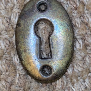 Könnte beinhalten: Ein antiker ovaler Schlüsselschlitzabdeckung aus Messing mit einer abgenutzten, gealterten Patina. Die Abdeckung hat zwei kleine Löcher oben und unten zum Befestigen.