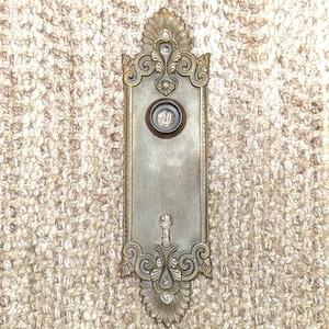 May include: A vintage brass door plate with an ornate design and a keyhole. The plate has a circular opening with a metal cover.