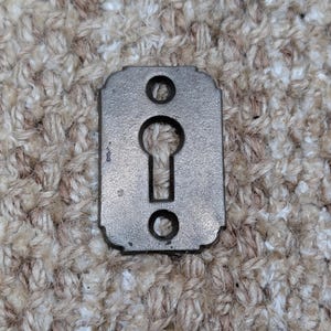 Könnte beinhalten: Eine rechteckige, antike Metall-Schlüssellochabdeckung mit dunkler, gealterter Oberfläche. Sie hat ein Schlüsselloch in der Mitte, zwei kreisförmige Löcher darüber und darunter sowie dekorative Kanten.