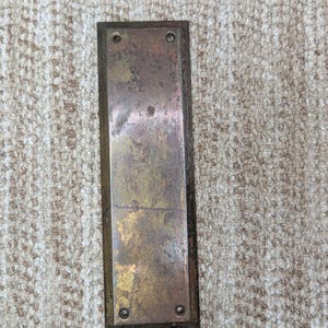 May include: A rectangular, weathered brass door push plate featuring a tarnished, mottled patina with visible signs of age and oxidation. The metal plate has four small, circular screw holes located at each of the four corners for mounting.