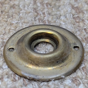 May include: A round, brass door knob escutcheon with a slightly raised edge and two small holes on the side. The escutcheon is worn and has a slightly tarnished finish.