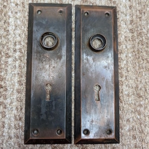 Op de afbeelding: Twee rechthoekige metalen deurplaten met een donkere, verweerde bronsafwerking. Elke plaat heeft een centrale ronde opening voor een deurknop, een sleutelgat eronder en vier kleine schroefgaten in de bovenste en onderste hoeken.